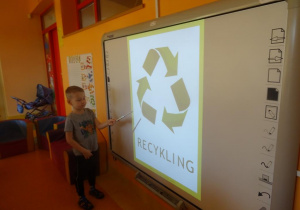 Chłopiec stoi pod tablicą interaktywną i wskaźnikiem pokazuje znak recyklingu.